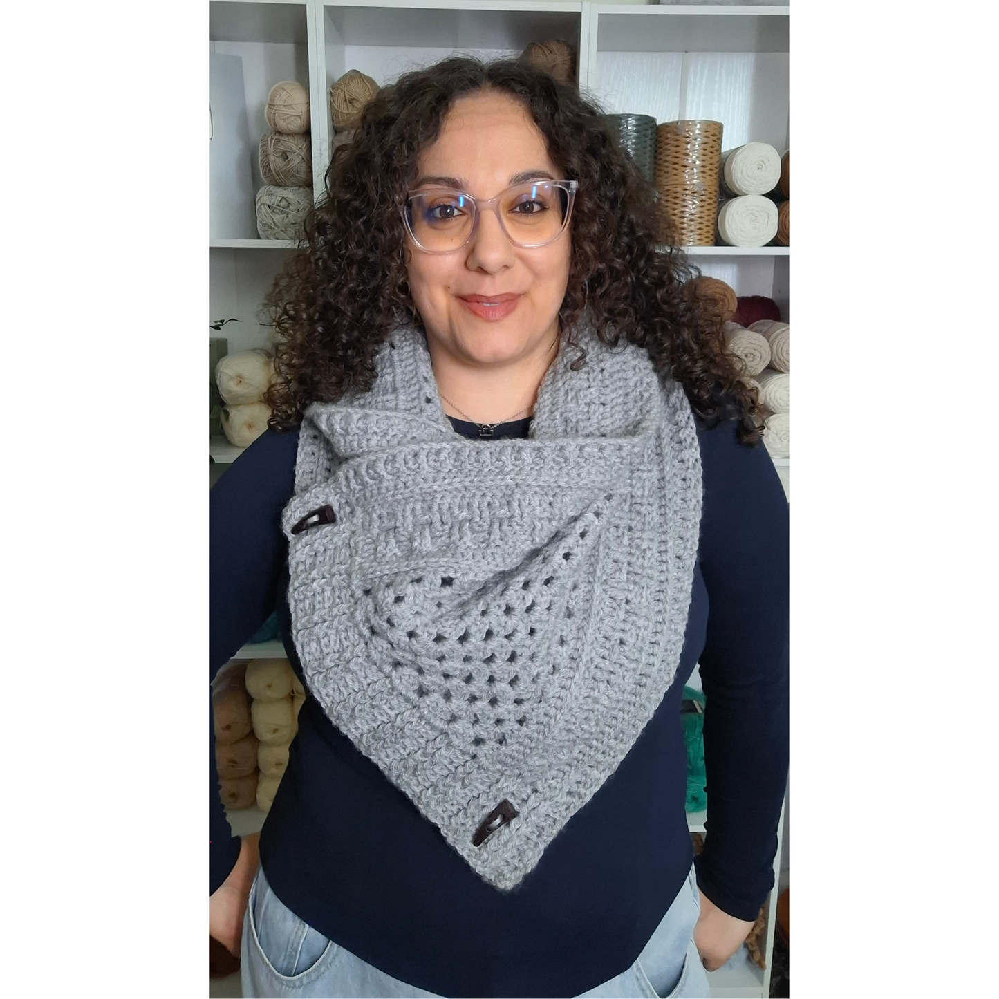 A woman with curly brown hair and glasses wearing a gray crocheted triangular cowl scarf with black buttons, standing in front of shelves of yarn.