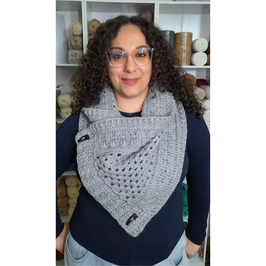 A woman with curly brown hair and glasses wearing a gray crocheted triangular cowl scarf with black buttons, standing in front of shelves of yarn.