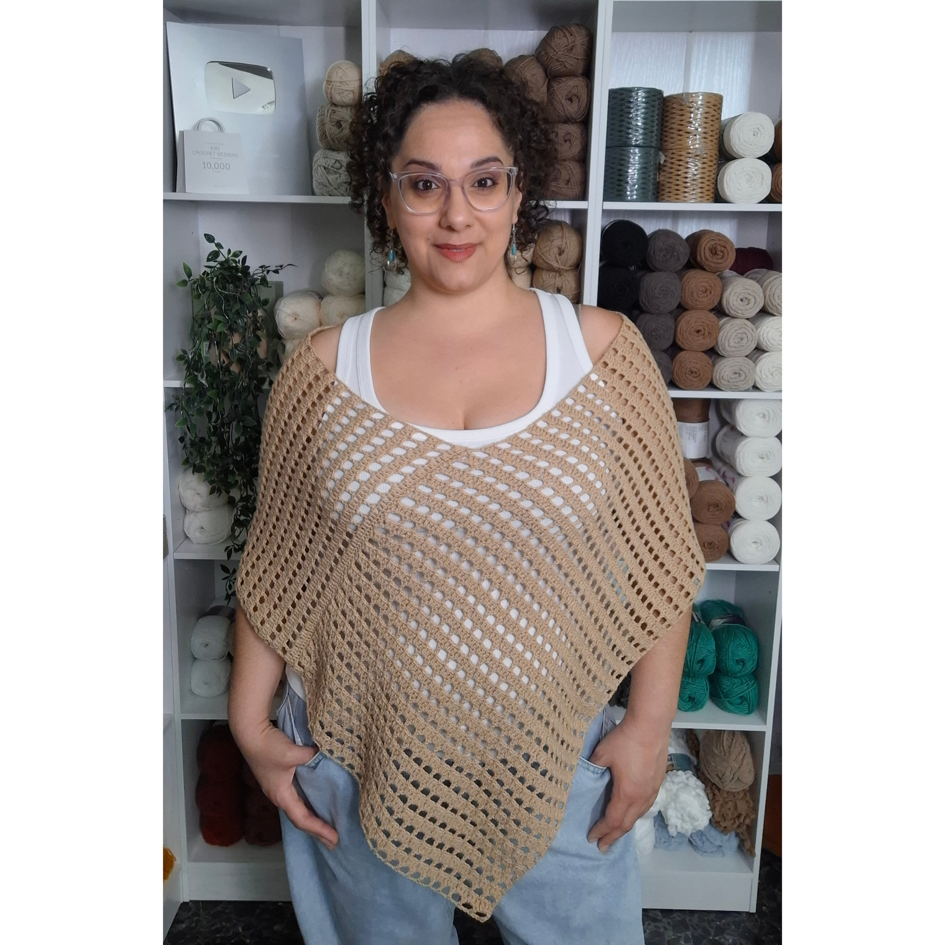 A woman wearing a beige open crocheted poncho over a white tank top and light blue jeans, standing in front of white shelves filled with yarn supplies.