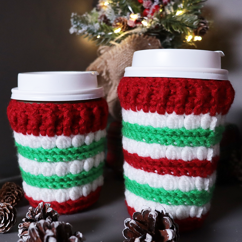 Two crocheted coffee cups with red, white, and green stripes in front of a Christmas tree.